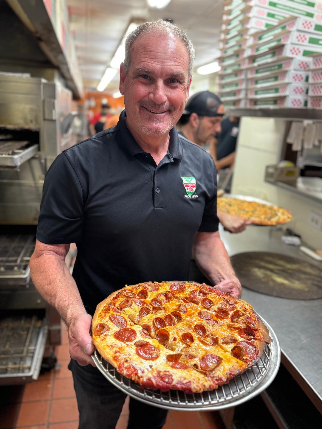 Joe Aurelio wearing a black polo with an Aurelio's logo holding a poperoni pizza in the restaurant